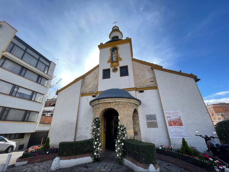 Parroquia Santa Monica fachada en tarde soleada con cielo azul, puerta decorada con flores. Al lado izquiero hay un edificio de apartamentos.