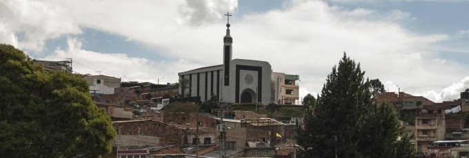 Santuario Nuestra Señora de la Peña vista de lejos en una cima detras de casas de barrio popular con cielo azul y nubes blancas. Destaca su fachada blanca con visos negros, su entrada en forma de óvalo y la torre con una punta alta que cierra con una cruz. 