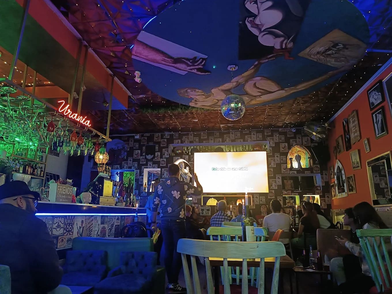  Brokeback Mountain interior del local decorado con fotos de colores, techo y paredes pintados. Personas conversando y viendo una pantalla.