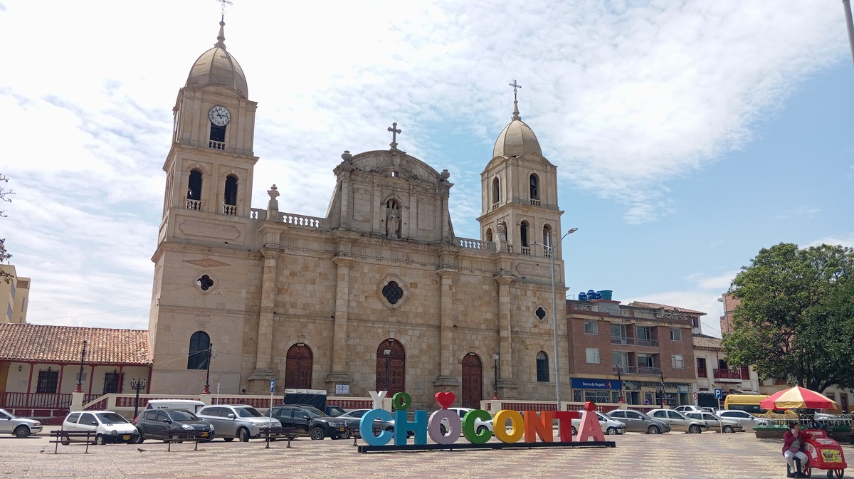  Iglesia Parroquial Nuestra Señora de la Salud fachada de frente de día con gigantografía con el nombre de Chocontá en la plaza rodeada de automóviles. Cielo azul con nubes blancas.