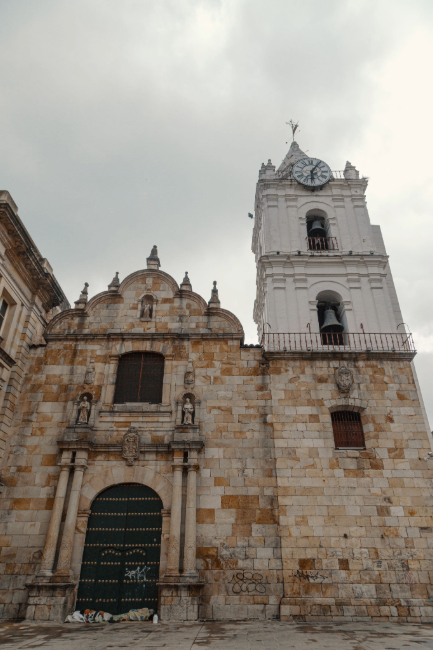  Iglesia de San Francisco fachada en primer plano en contrapicado, estructura entre piedra y concreto con puerta en madera ovalada. Destaca la superficie blanca de la torre del reloj y campanario.