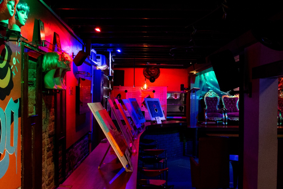  Museo LGBTI sala oscura con paredes con adornos de colores y fotografías. Se ven 3 cabezas de maniquies y dos sillas clásicas.