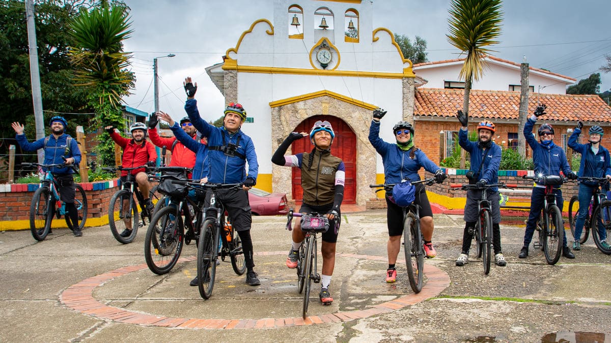 Grupo de ciclistas al frente de una iglesia en pueblo, saludan a un dron.