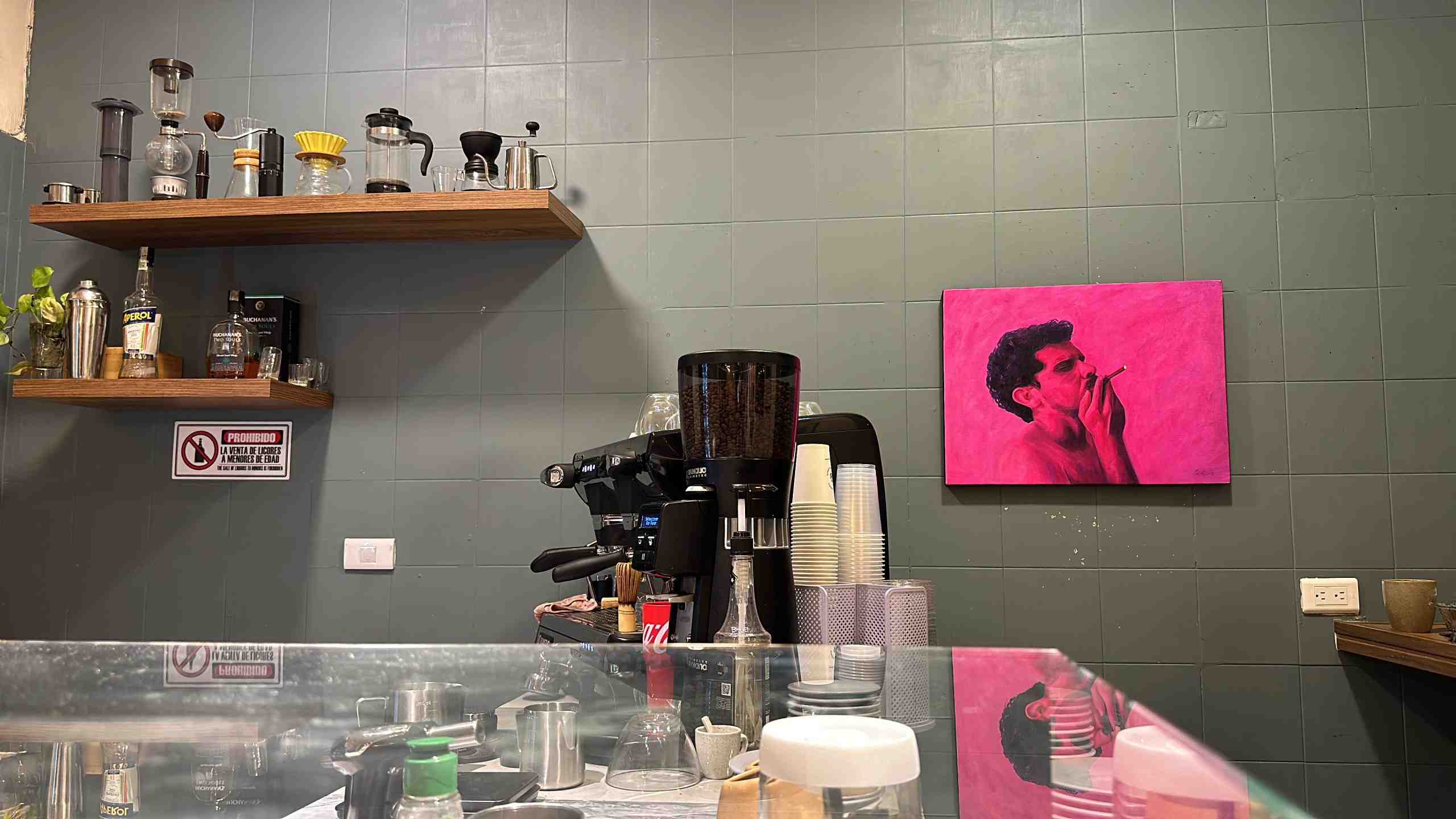 Interior de una cocina con accesorios para preparar café. Hay una pared gris al fondo con el cuadro de un hombre en color rosado y que está fumando.
