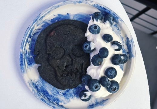 Postre con galleta, helado y arándanos. La galleta es negra y tieje forma de calabera. El postre está sobre un plato blanco con manchas azules.