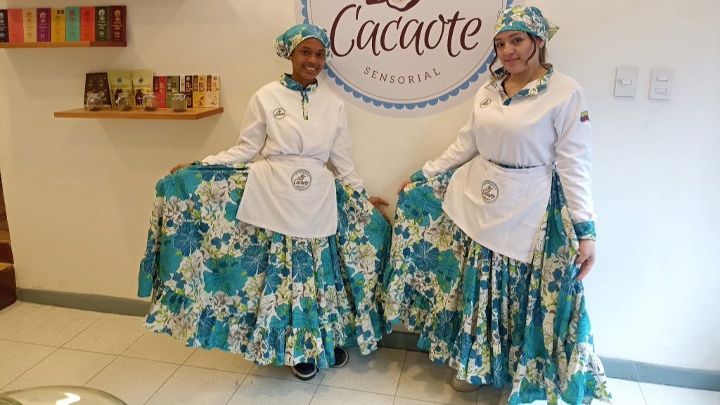 Dos mujeres jóvenes vestidas con delantales de color azul y blanco donde resaltan varias leyendas que dicen "Cacaote"