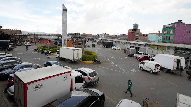 Corabastos, plano general de una gran plazoleta, rodeada de locales comerciales. Hay varios caminones alrededor. Hay un obelisco blanco en la mitad.