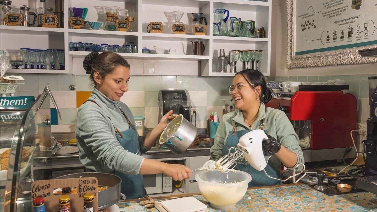 Un par de mujeres jóvenes en una cocina preparan y mezclan algo en un platón de plástico. Están sonriendo.