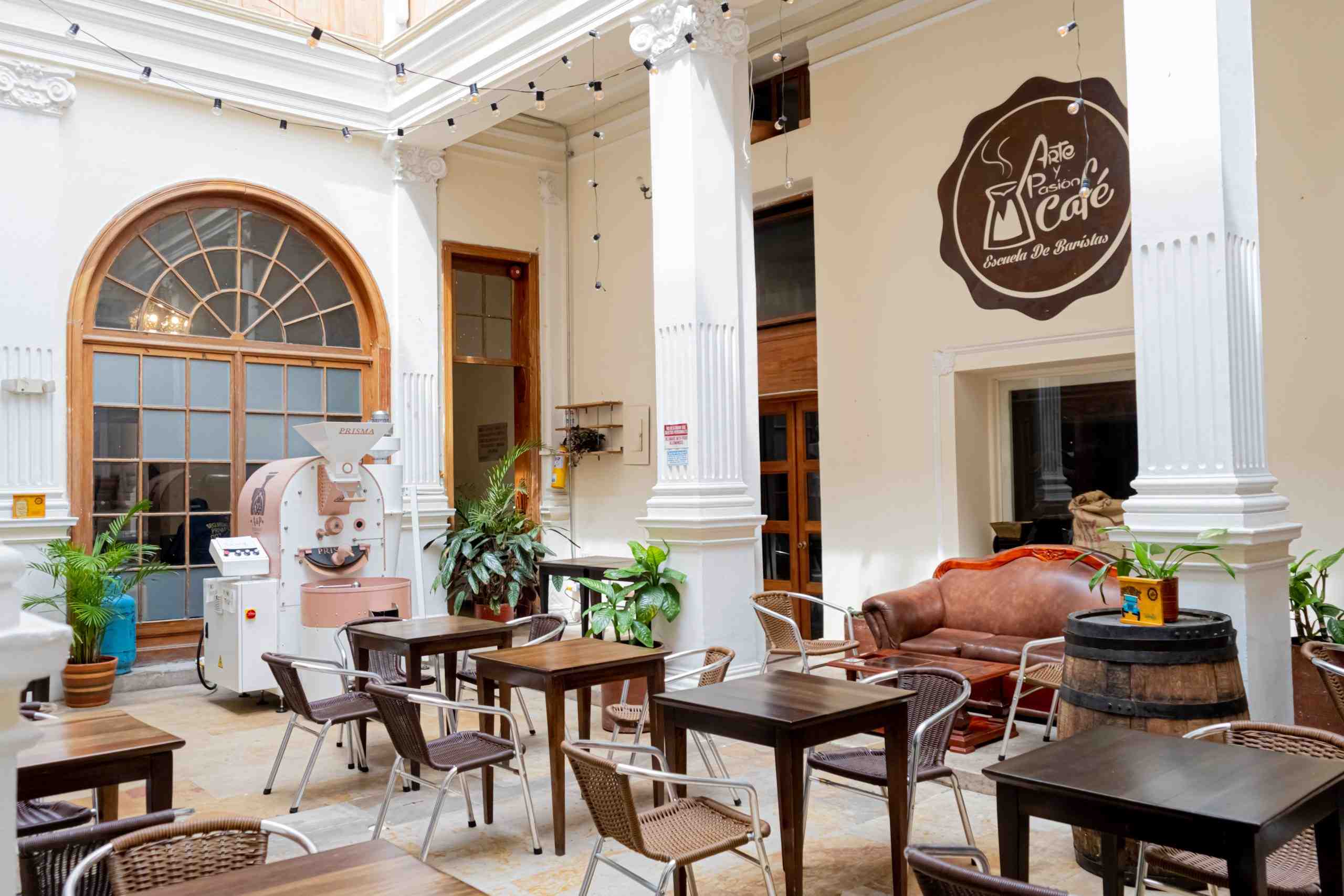 Salón principal de un café en el que se ven mesas de madera y sillas de mimbre, 3 columnas de mármol que sostienen el lugar y le dan un elegante decorado y un aviso en una pared que dice "Arte y pasión café".