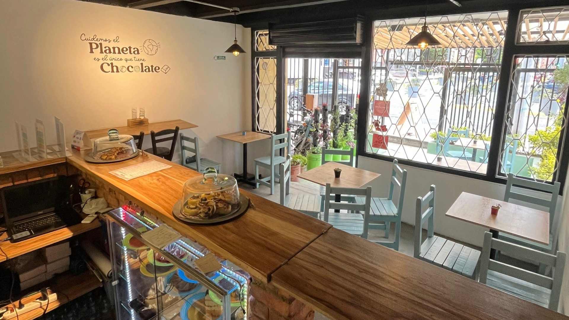 Grupo de personas jóvenes alrededor de una mesa escuchan a un mesero que viste con un delantal y una gorra azul. 