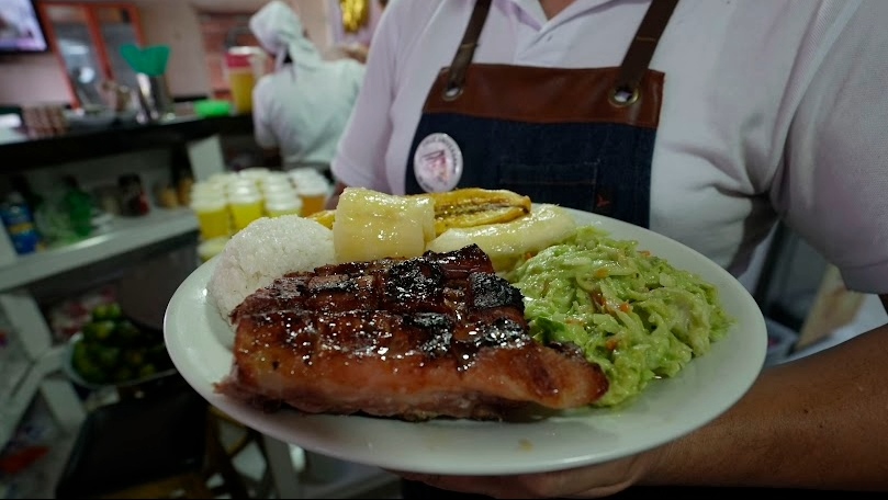 Primer plano de un plato de comida sostenido por un mesero en el que se ve un gran pedazo de carne asada, arroz blanco, yuca, plátano y ensalada de aguacate.