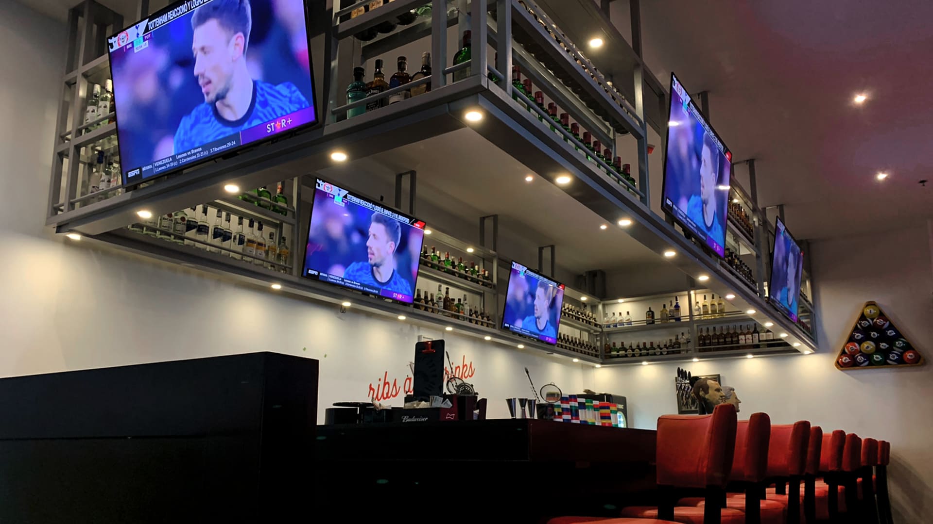 Un bar deportivo moderno con múltiples televisores transmitiendo partidos, una barra bien surtida y sillas rojas.