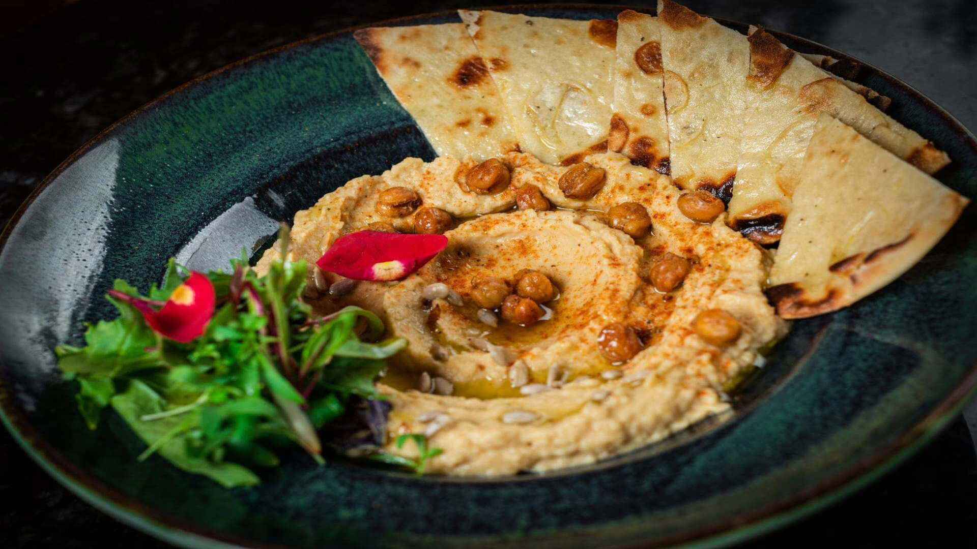 Hummus cremoso con garbanzos, semillas de girasol, pimentón y pan de pita, servido en un plato verde.