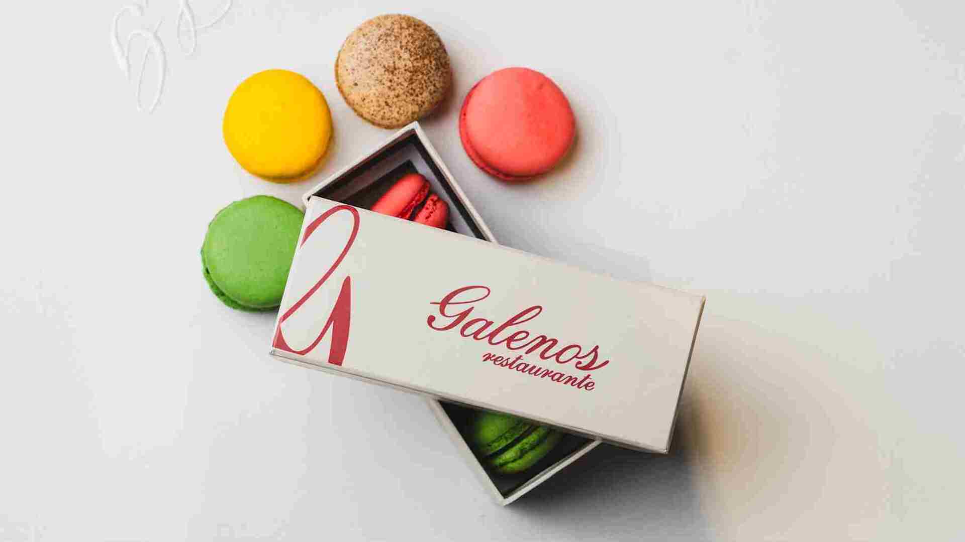 Varios macarons de colores vivos (verde, amarillo, rosa, marrón) esparcidos alrededor de una caja abierta de "Galenos restaurante".