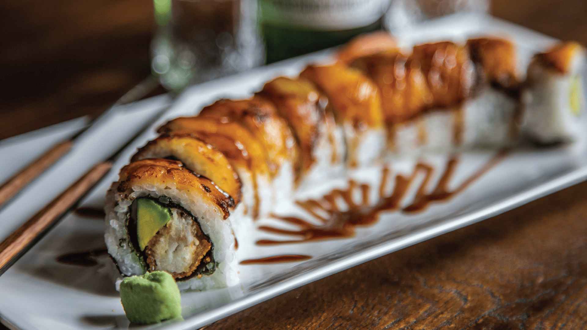Un rollo de sushi generosamente bañado en salsa, con aguacate y un relleno crujiente, servido con wasabi en un plato blanco.
