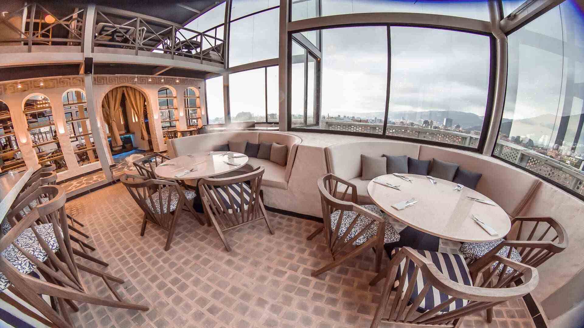 Un elegante restaurante en la azotea con ventanales de piso a techo, ofreciendo vistas panorámicas de la ciudad.