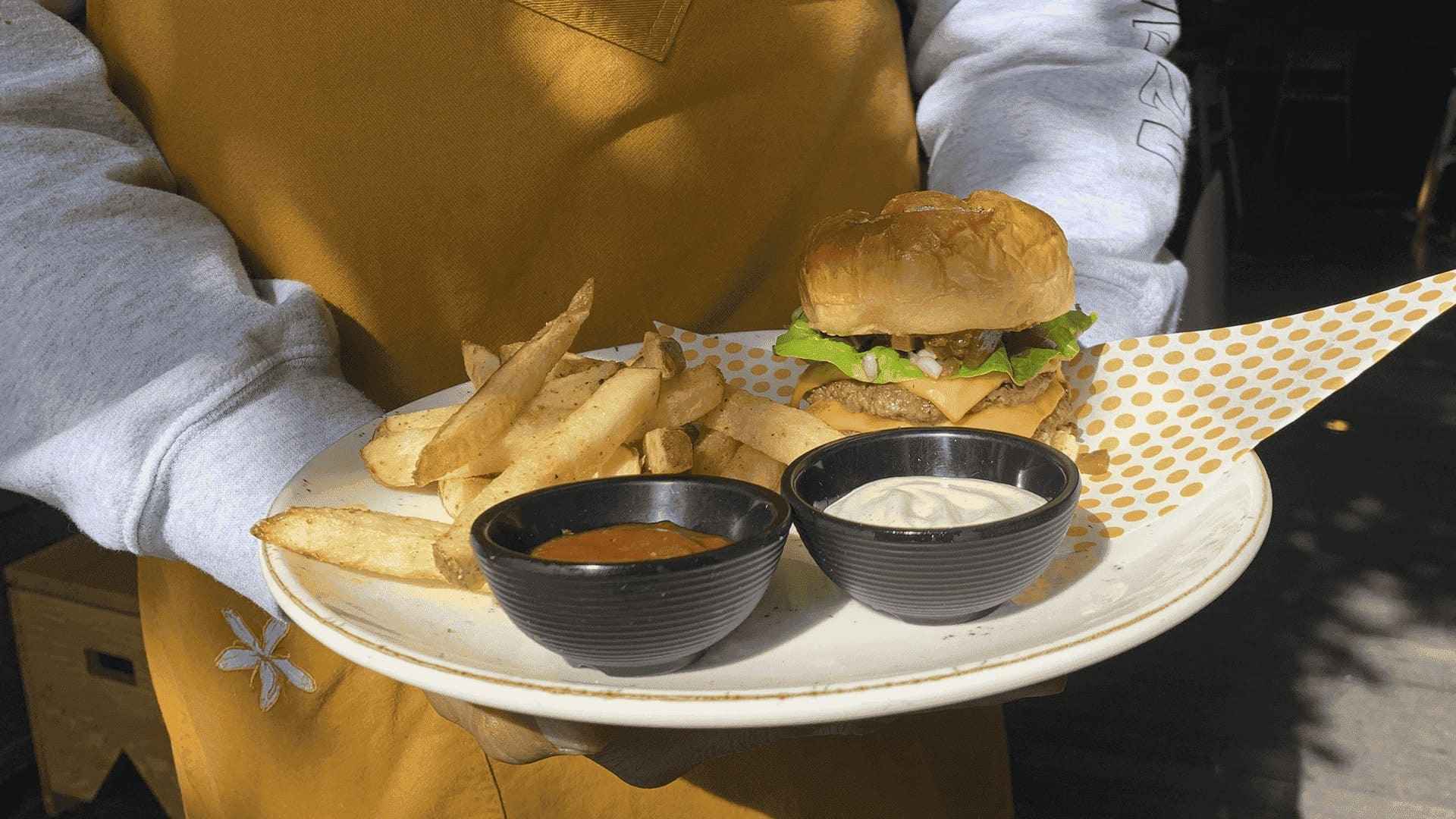 Hamburguesa con papas fritas, kétchup y mayonesa en recipientes negros, servida en plato blanco.
