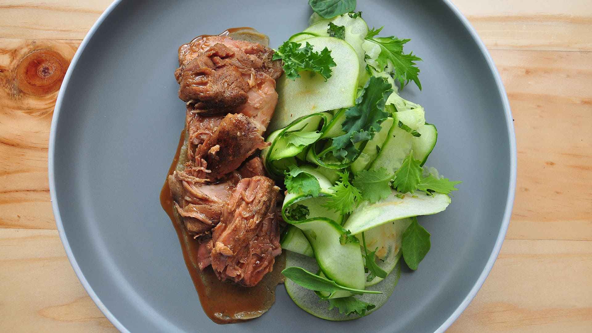 Un plato con carne desmenuzada en salsa y una ensalada fresca de pepino y hojas verdes.