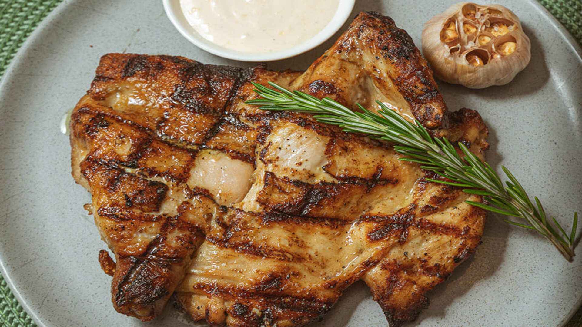 Una pechuga de pollo a la parrilla con marcas de la parrilla, romero fresco y una cabeza de ajo asada.