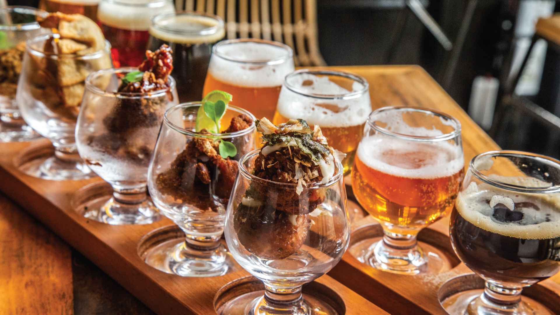 Una degustación variada de cervezas artesanales y aperitivos en vasos pequeños, sobre una bandeja de madera, invitando a probar nuevos sabores.