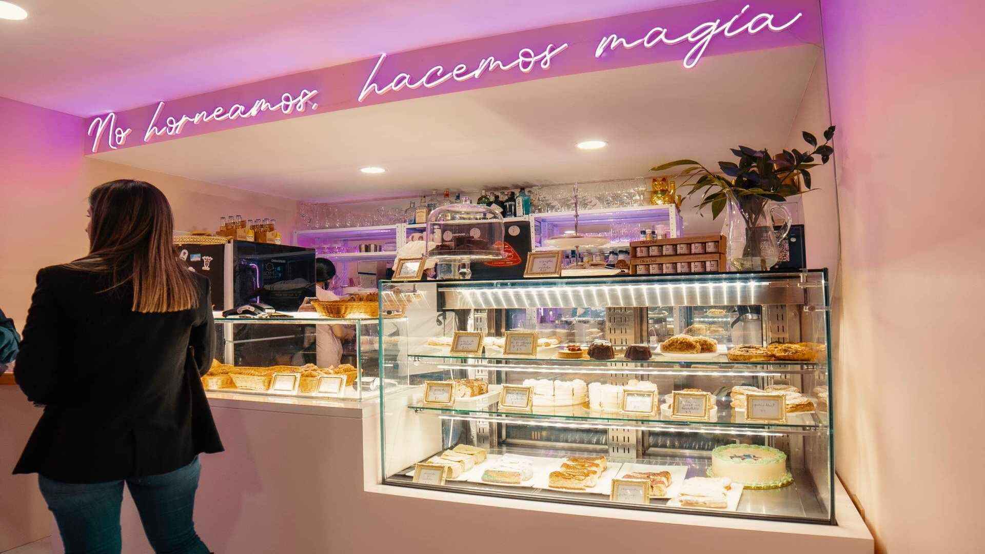 El interior de una pastelería moderna con una exhibición de postres y un letrero de neón que dice "No horneamos, hacemos magia".