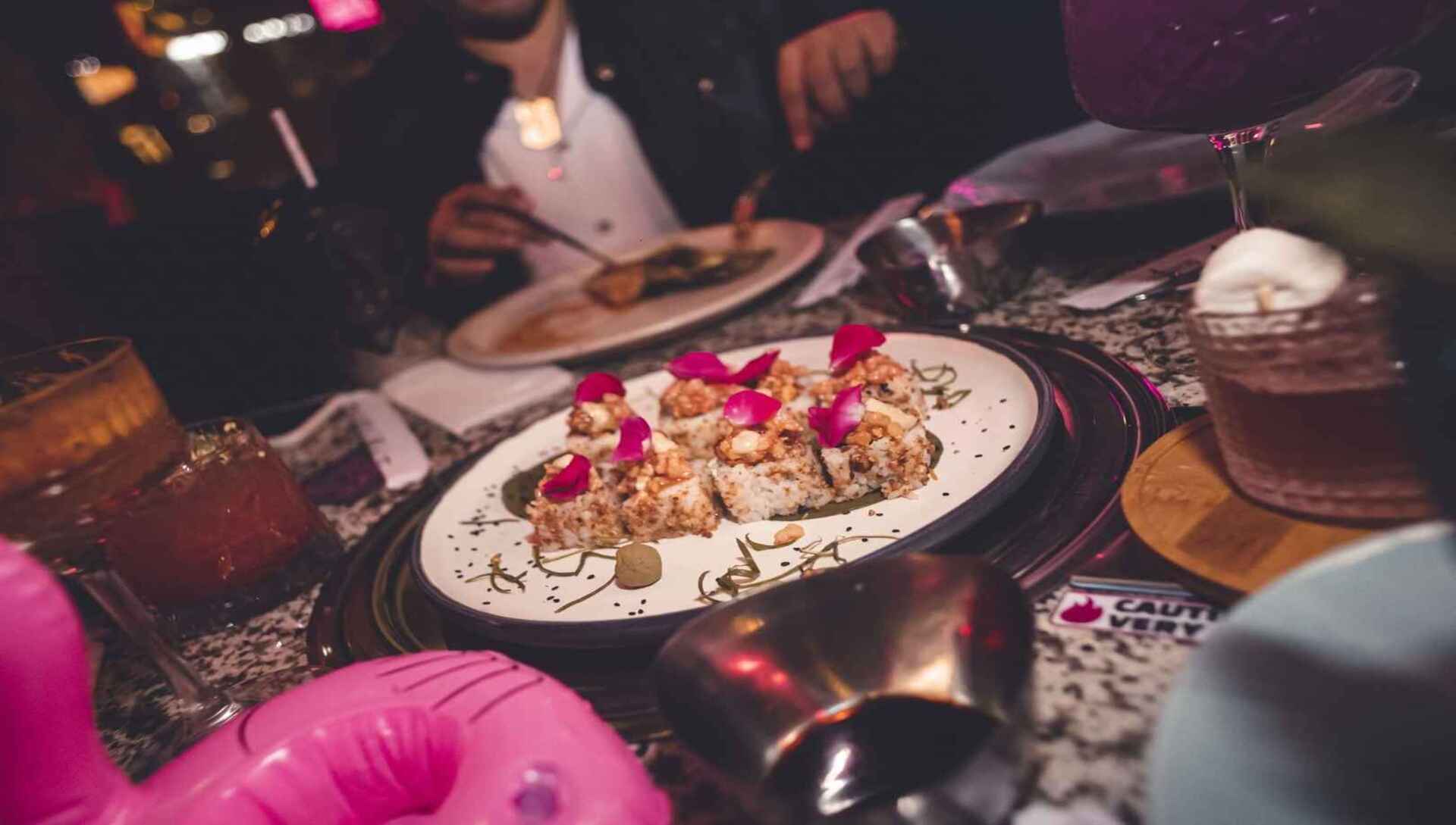 Rollos de sushi con pétalos rosados, servidos en una mesa con cócteles y decoración lúdica de flamenco.