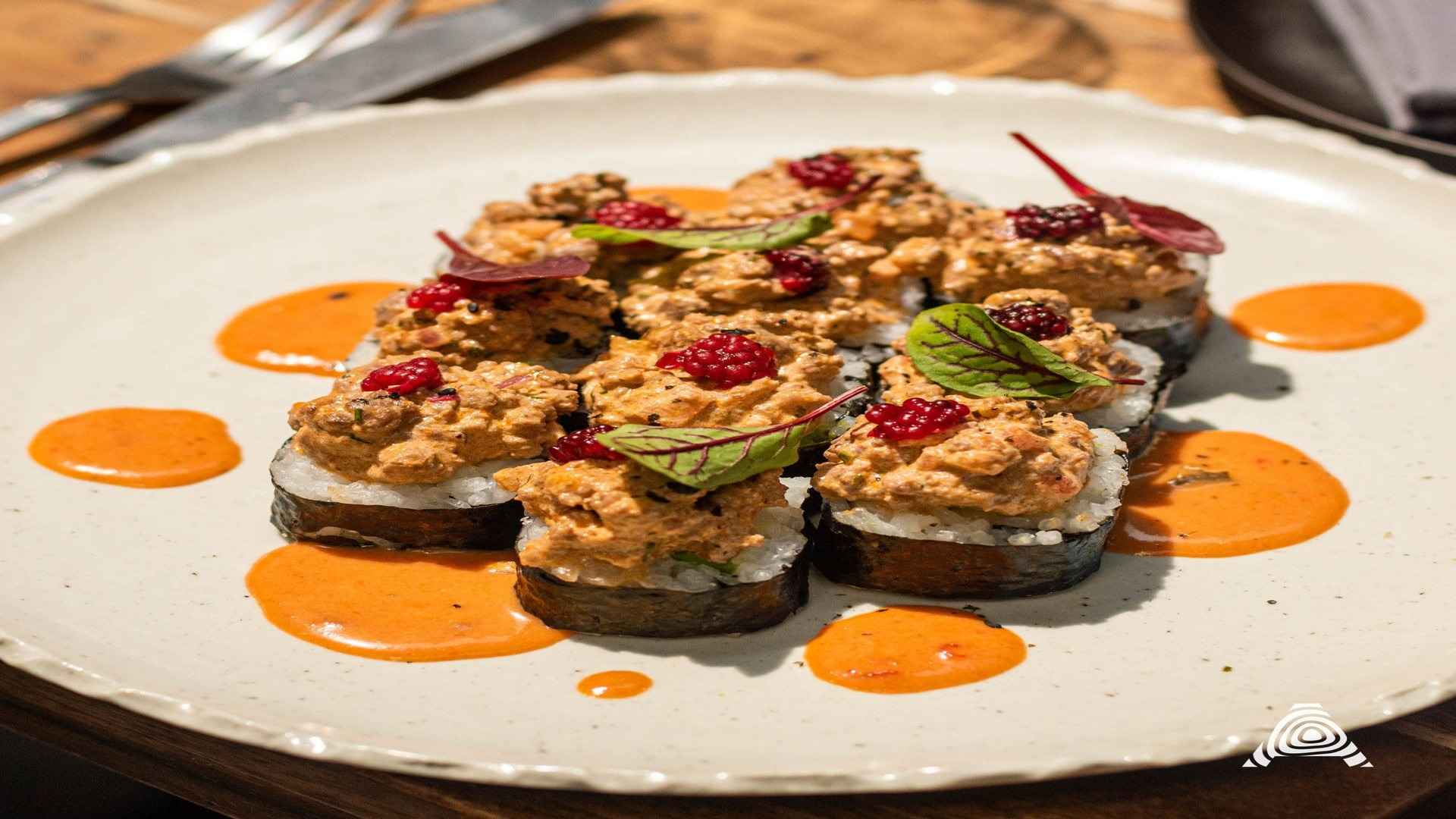 Rollos de sushi creativamente emplatados con una cobertura generosa y vibrantes bayas rojas, acompañados de salsa naranja.