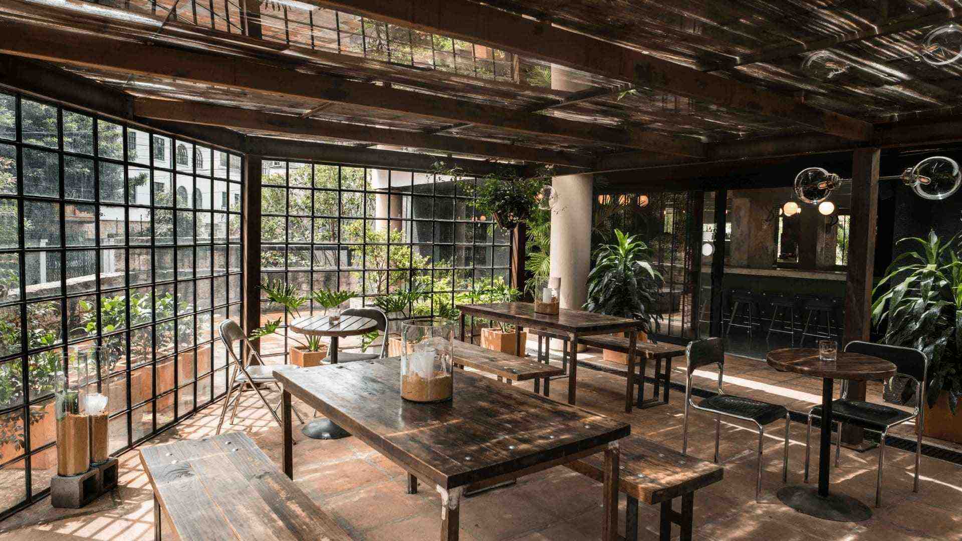 Un comedor de restaurante con techo de cristal, mesas y sillas de madera, y plantas verdes.