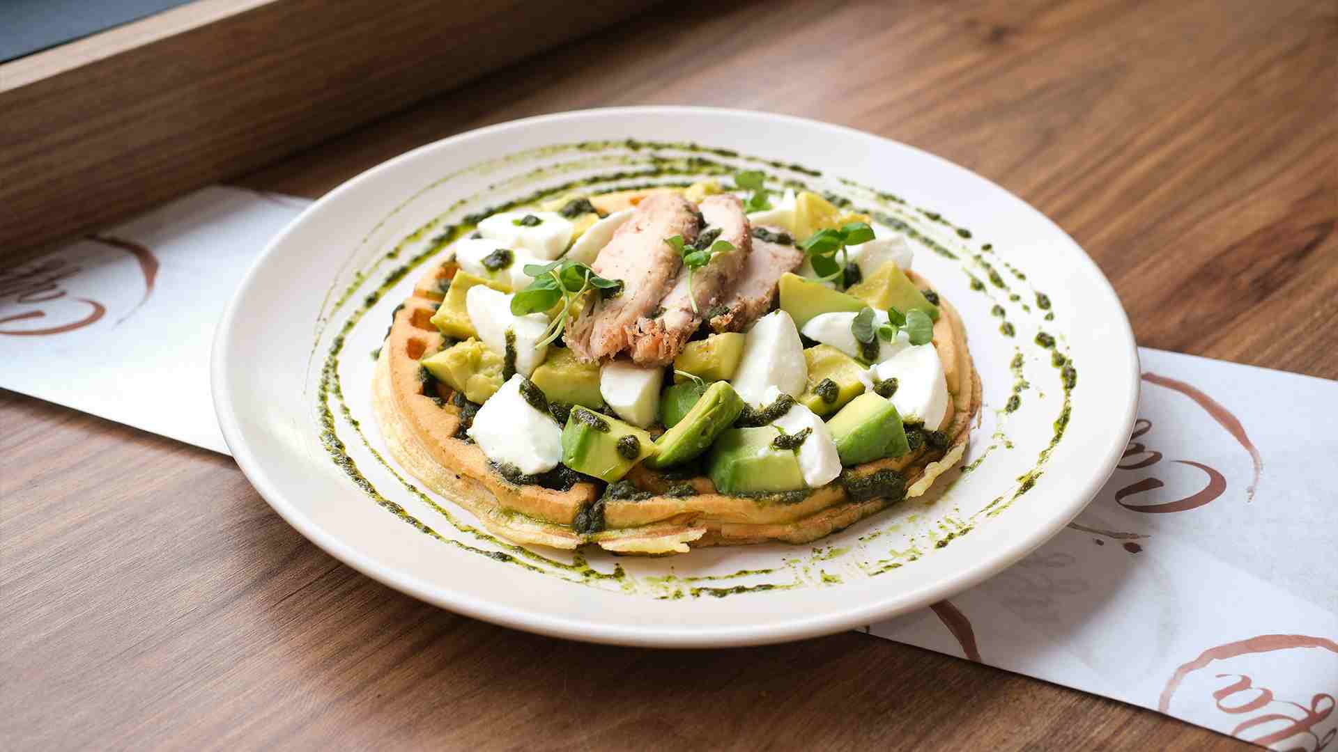 Un gofre salado gourmet con aguacate, queso mozzarella, pollo y pesto, servido en un plato blanco.