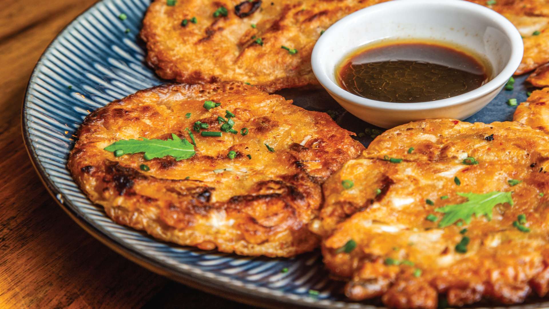 Tres pequeñas tortitas doradas y crujientes, con hierbas frescas, acompañadas de una salsa oscura en un cuenco blanco.