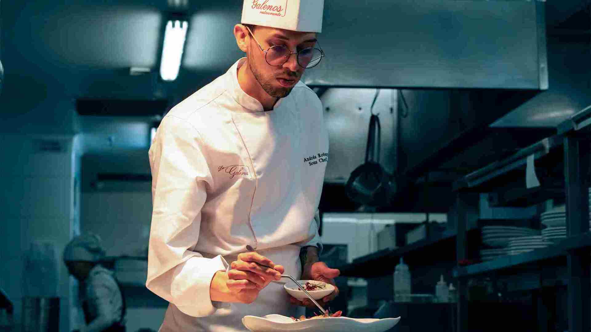 Un chef concentrado, con gafas y uniforme blanco, emplata meticulosamente un plato en una cocina profesional.