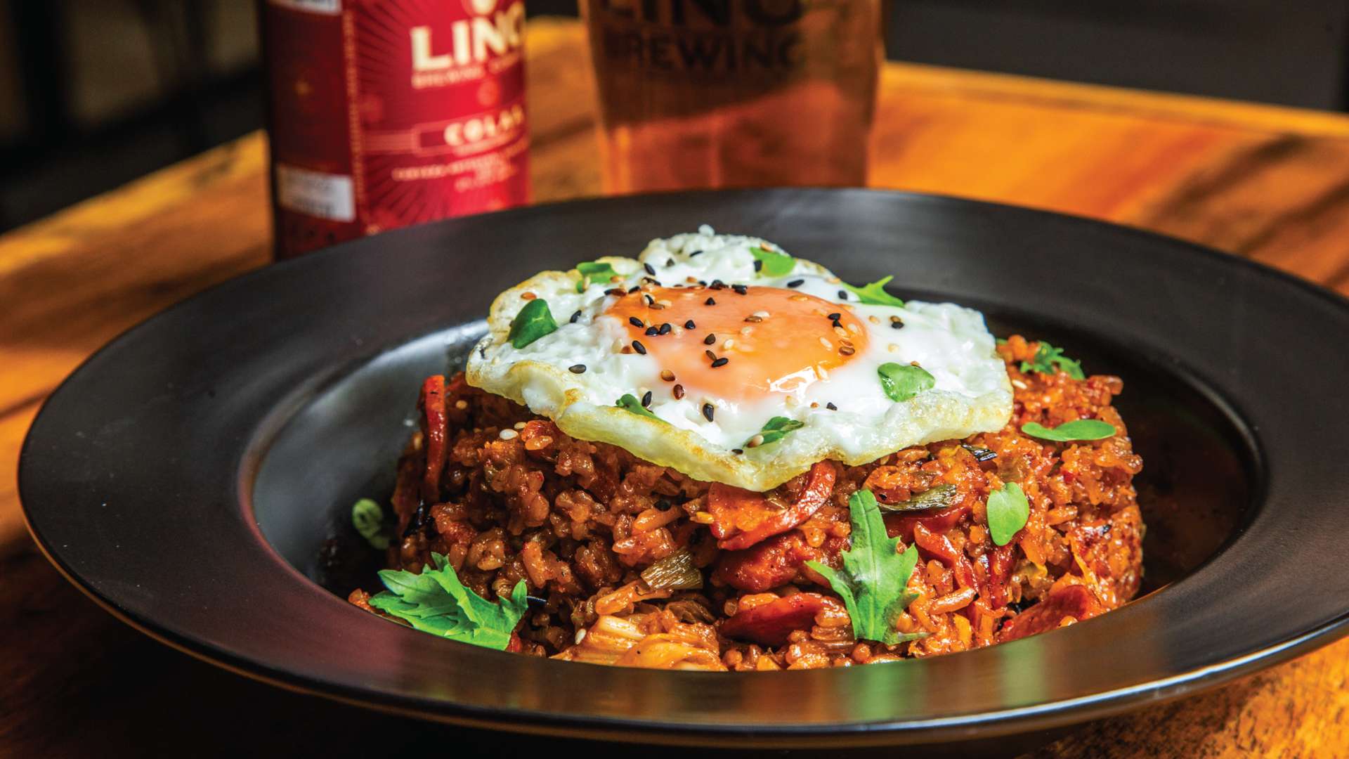 Un plato de arroz frito picante con kimchi, coronado con un huevo frito y espolvoreado con semillas de sésamo, con una bebida de fondo.