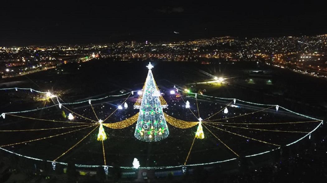  Un gran árbol de Navidad iluminado de noche. Al fondo se ve la ciudad también iluminada.