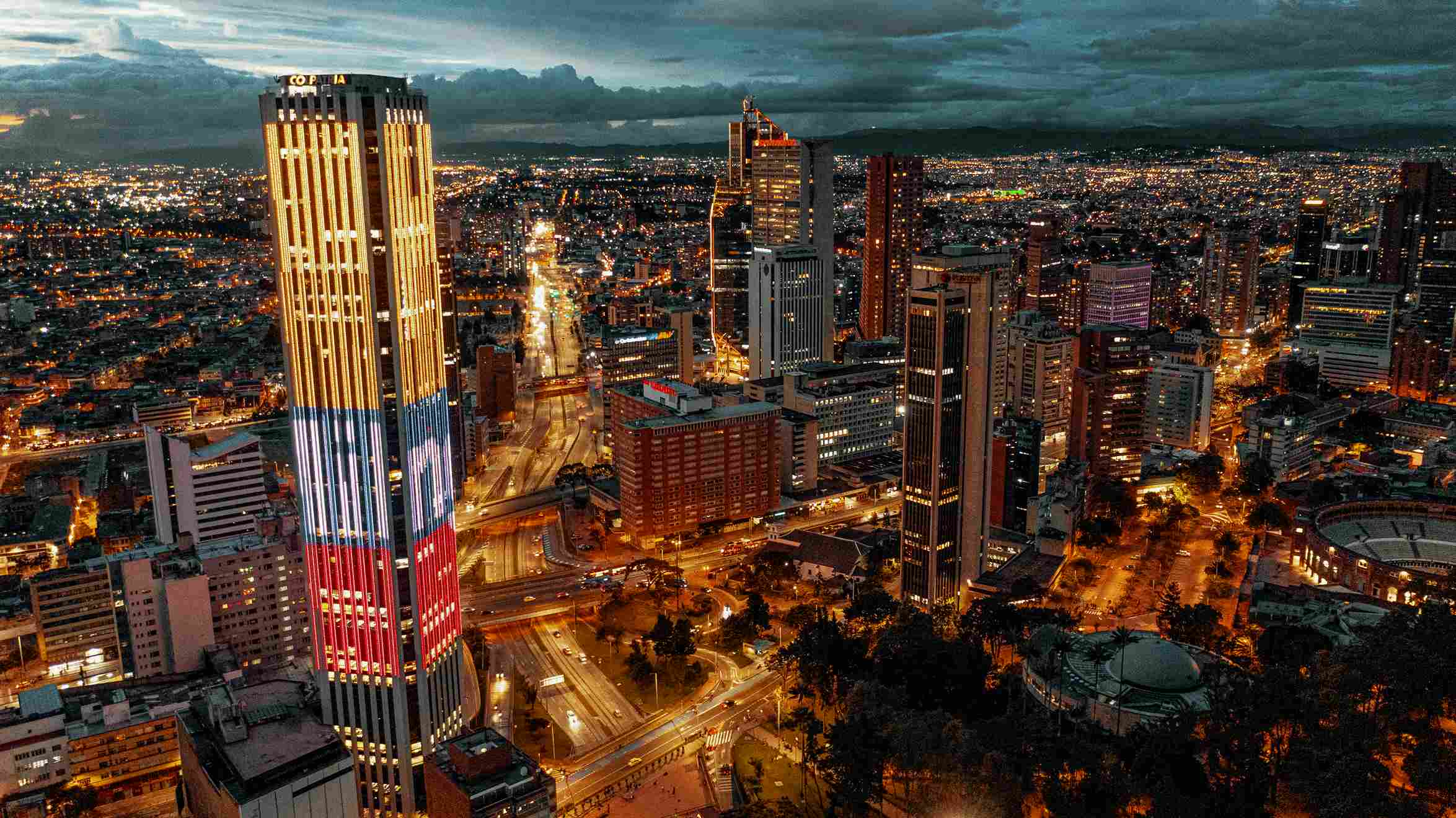  Panorámica de Bogotá en la que se observa en primer plano la Torre Colpatria iluminada con los colores de la bandera de Colombia, la calle 26 hacia el occidente y una serie de edificios hacia el occidente.