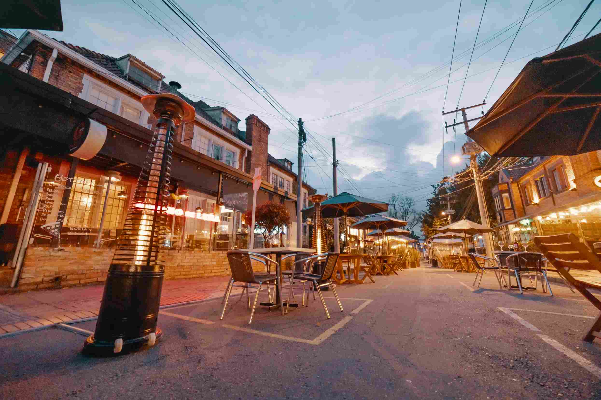  Una calle al atardecer con mesas alrededor cubiertas por carpas. Cada una tiene un calentador de ambiente de gas en forma de pirámide.