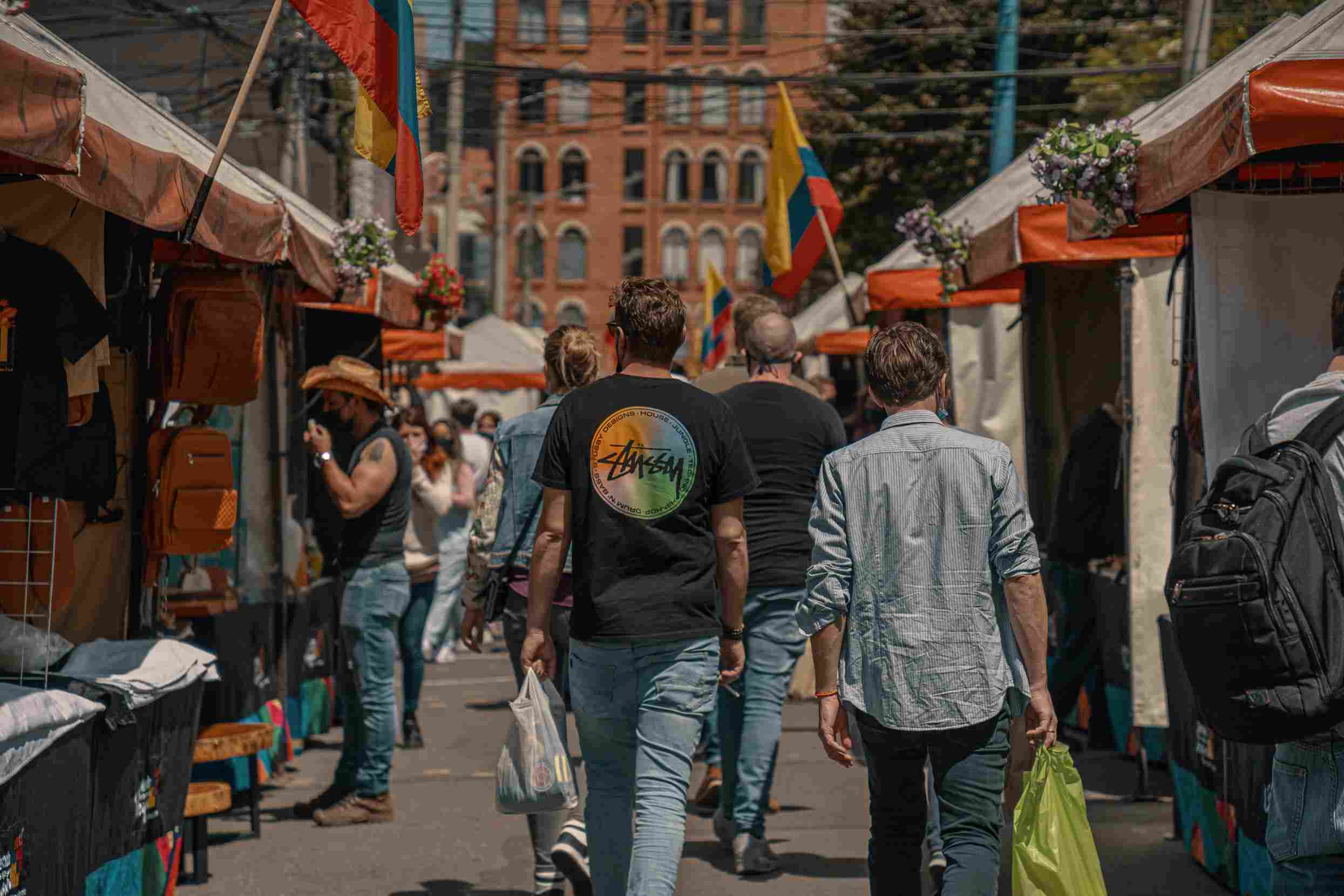 Hombres jóvenes, parecen turistas, que caminan al frente de unos toldos de comercio callejero. En los lugares se observan varias banderas de Colombia.