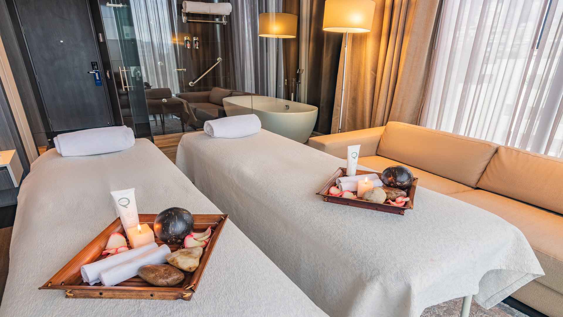 Dos camillas de masaje preparadas en una suite de hotel con una bañera independiente al fondo. El ambiente es lujoso y relajante, ideal para tratamientos de spa privados.