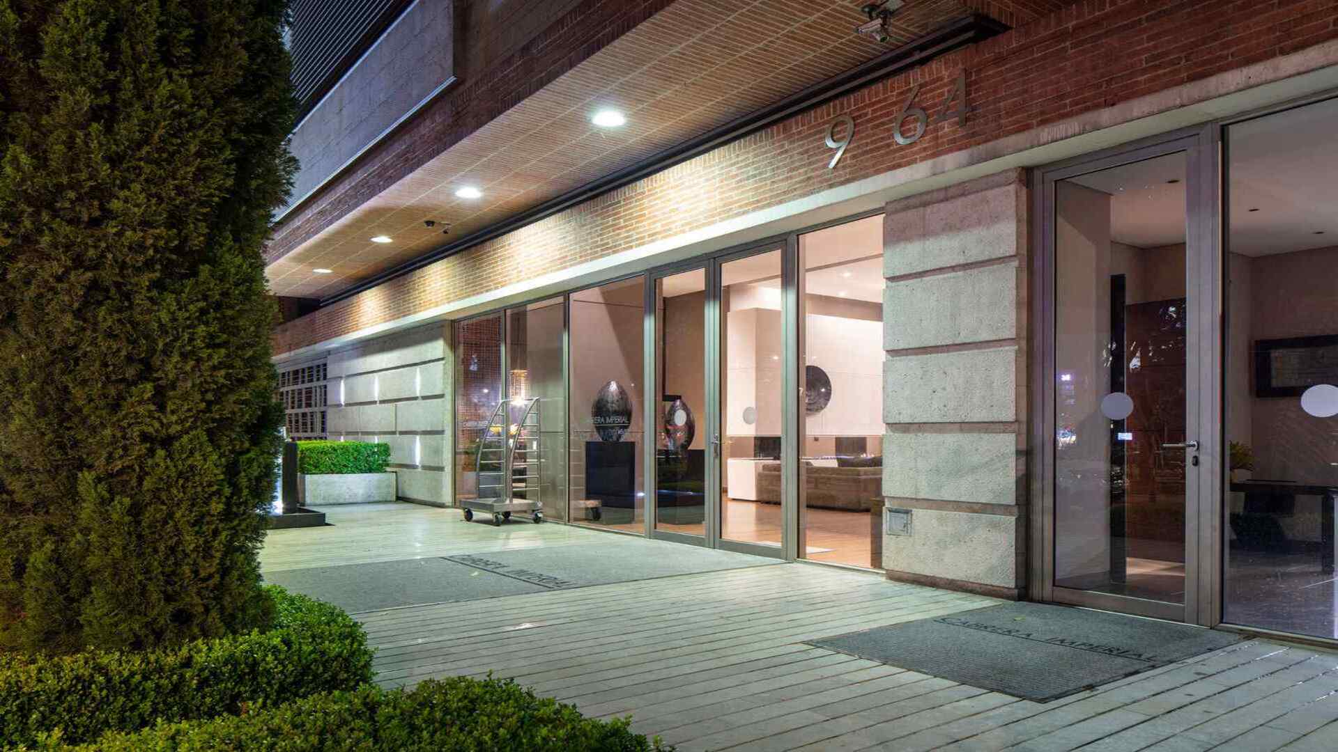 Entrada moderna de un edificio con puertas de cristal y ladrillo a la vista. Un carrito de equipaje espera, y se insinúa un lobby elegante en el interior.