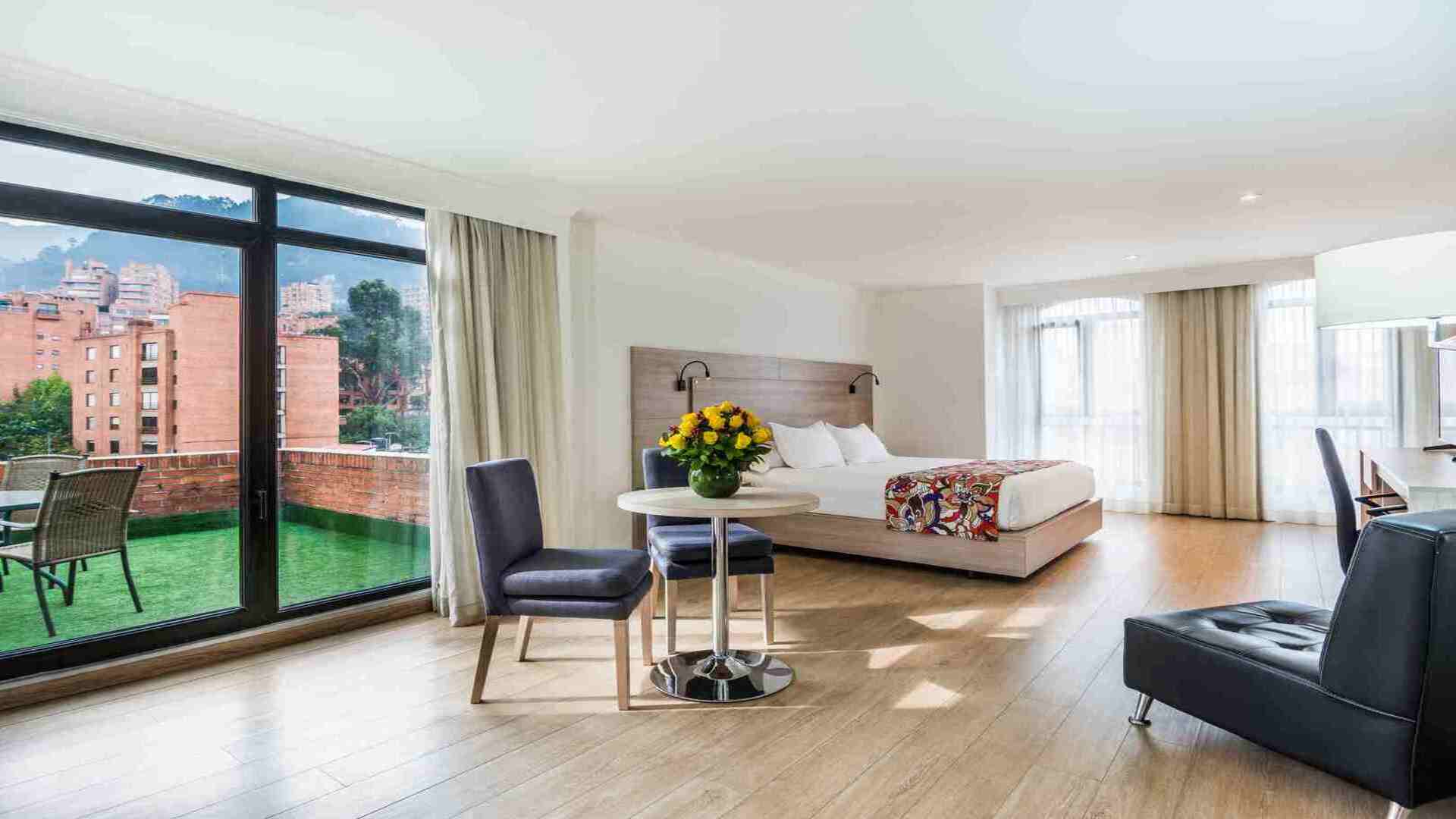 Una suite espaciosa con una cama grande, zona de estar, y un balcón exterior con césped artificial y muebles. Grandes ventanales ofrecen vistas