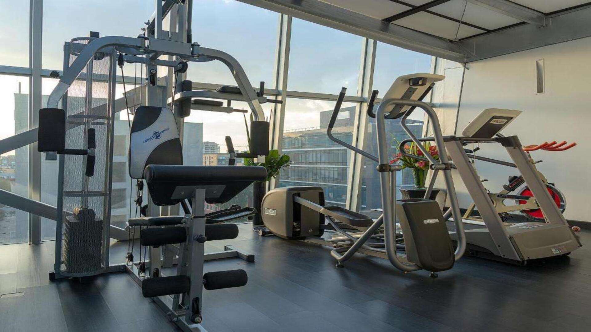 Gimnasio de hotel con máquinas de ejercicio y grandes ventanales con vistas urbanas.