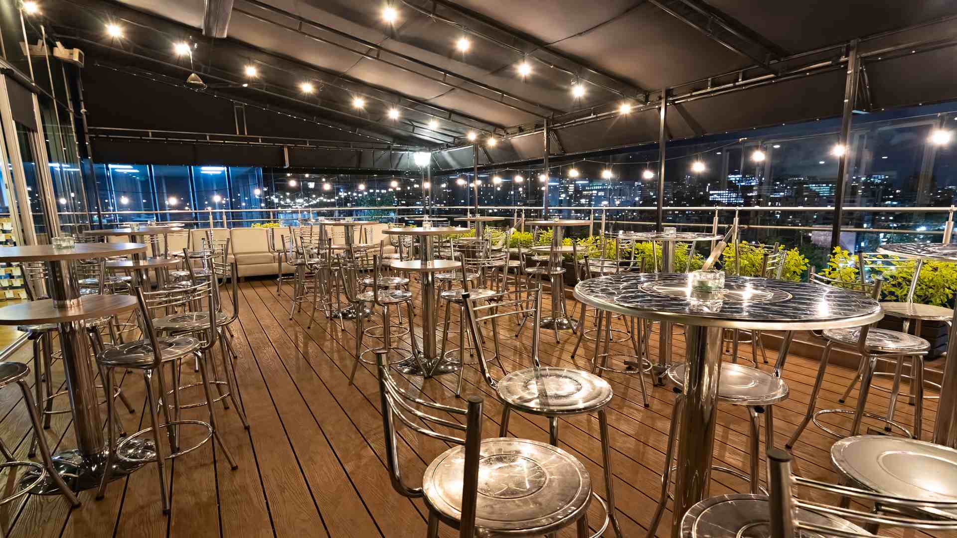  Una terraza de noche con mesas y sillas altas de metal, luces colgantes y vistas a la ciudad. Un ambiente vibrante para socializar.