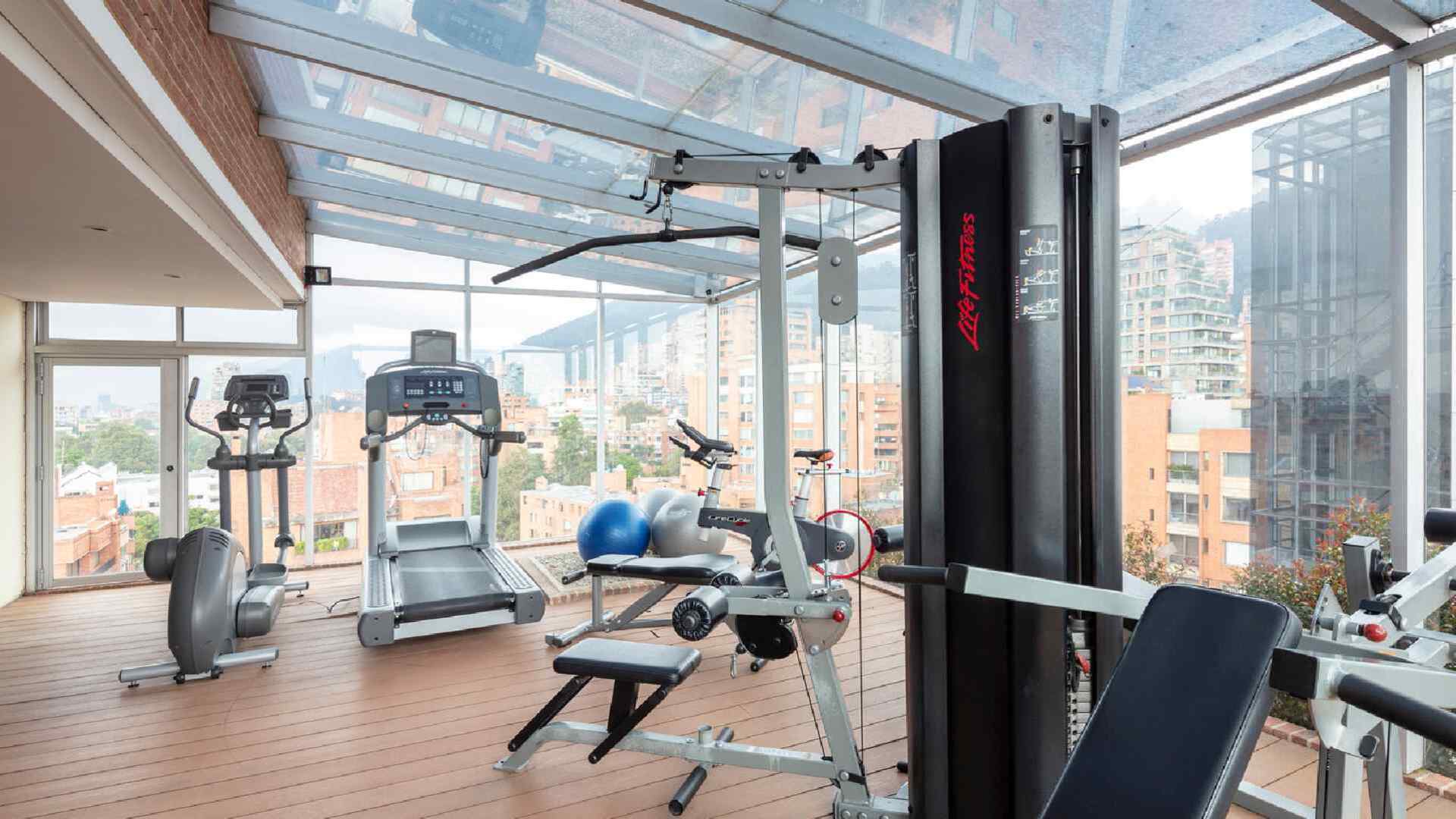 Un gimnasio en la azotea con techo de cristal que ofrece vistas panorámicas de la ciudad. Equipado con diversas máquinas de ejercicio para un entrenamiento con paisaje.