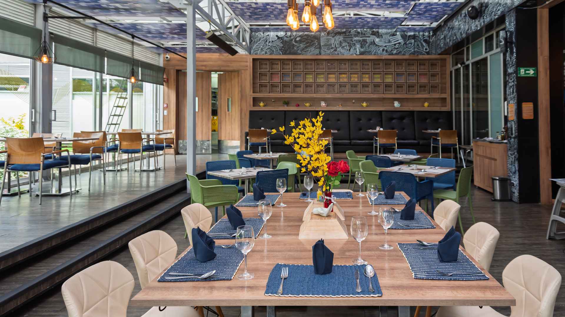 Un restaurante elegante y moderno con mesas dispuestas para cenar, sillas de colores, y una decoración única con un mural y estanterías de madera.