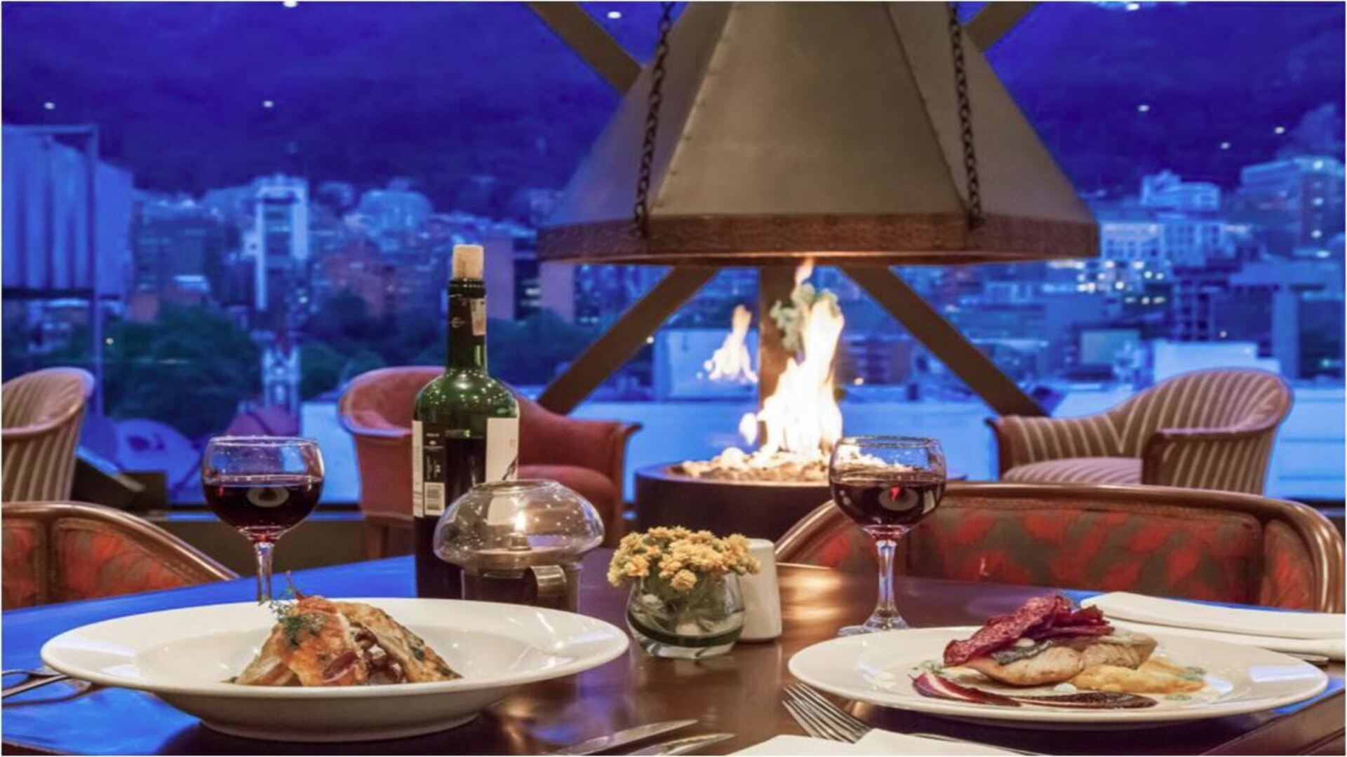  Una cena romántica en un restaurante con vistas a la ciudad, con un ambiente cálido creado por una chimenea central. Se aprecian platos gourmet y copas de vino.