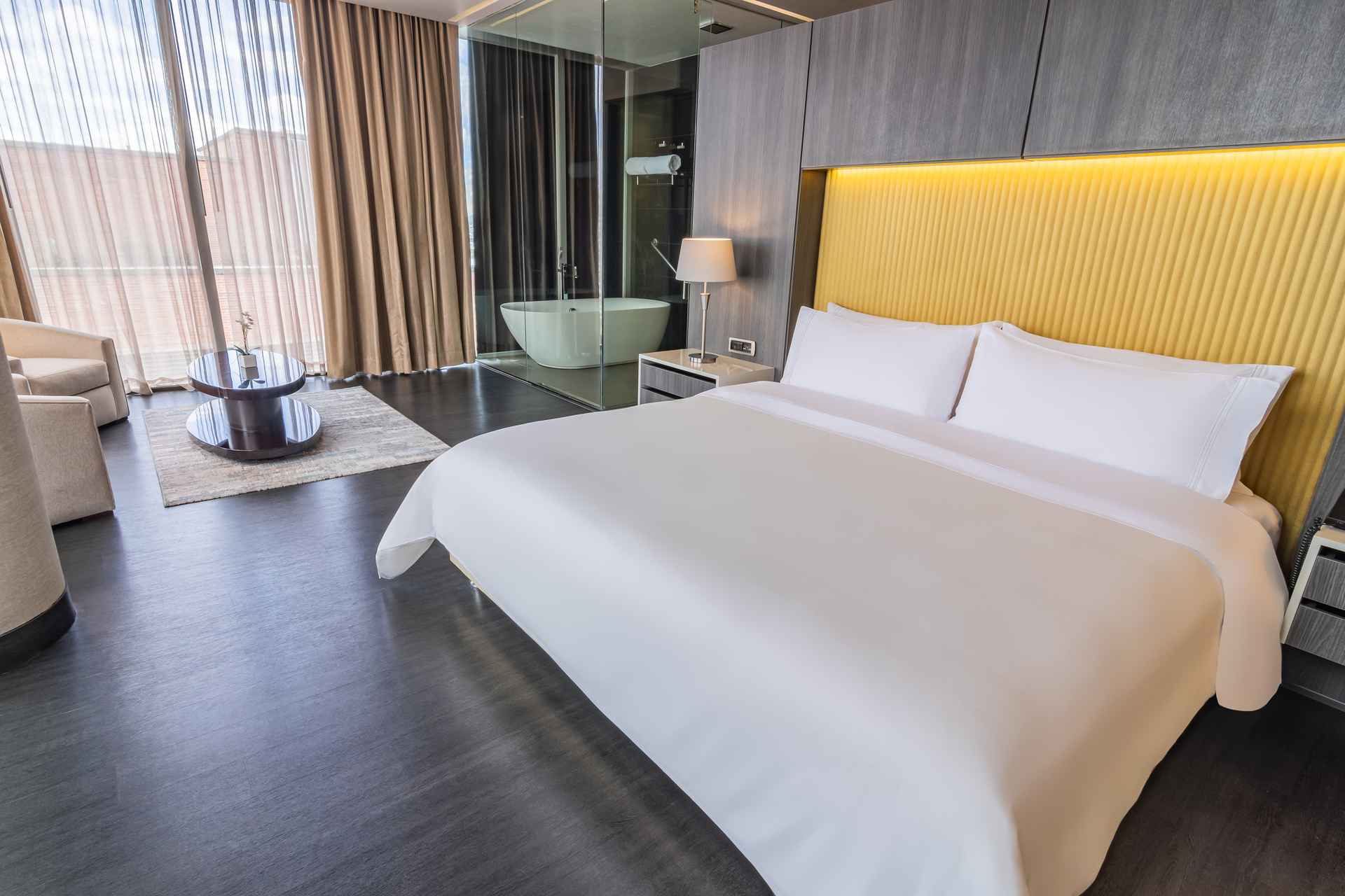 Una lujosa habitación de hotel con una cama king-size, cabecera amarilla, y un baño con bañera independiente visible a través de un panel de cristal.