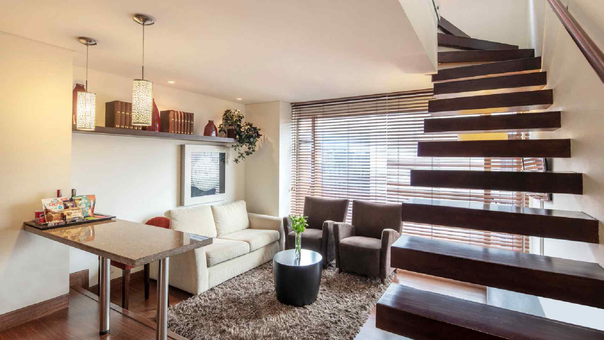  Una sala de estar en un apartamento tipo dúplex, con un sofá, una zona de bar y una escalera de madera flotante que conduce al segundo nivel.