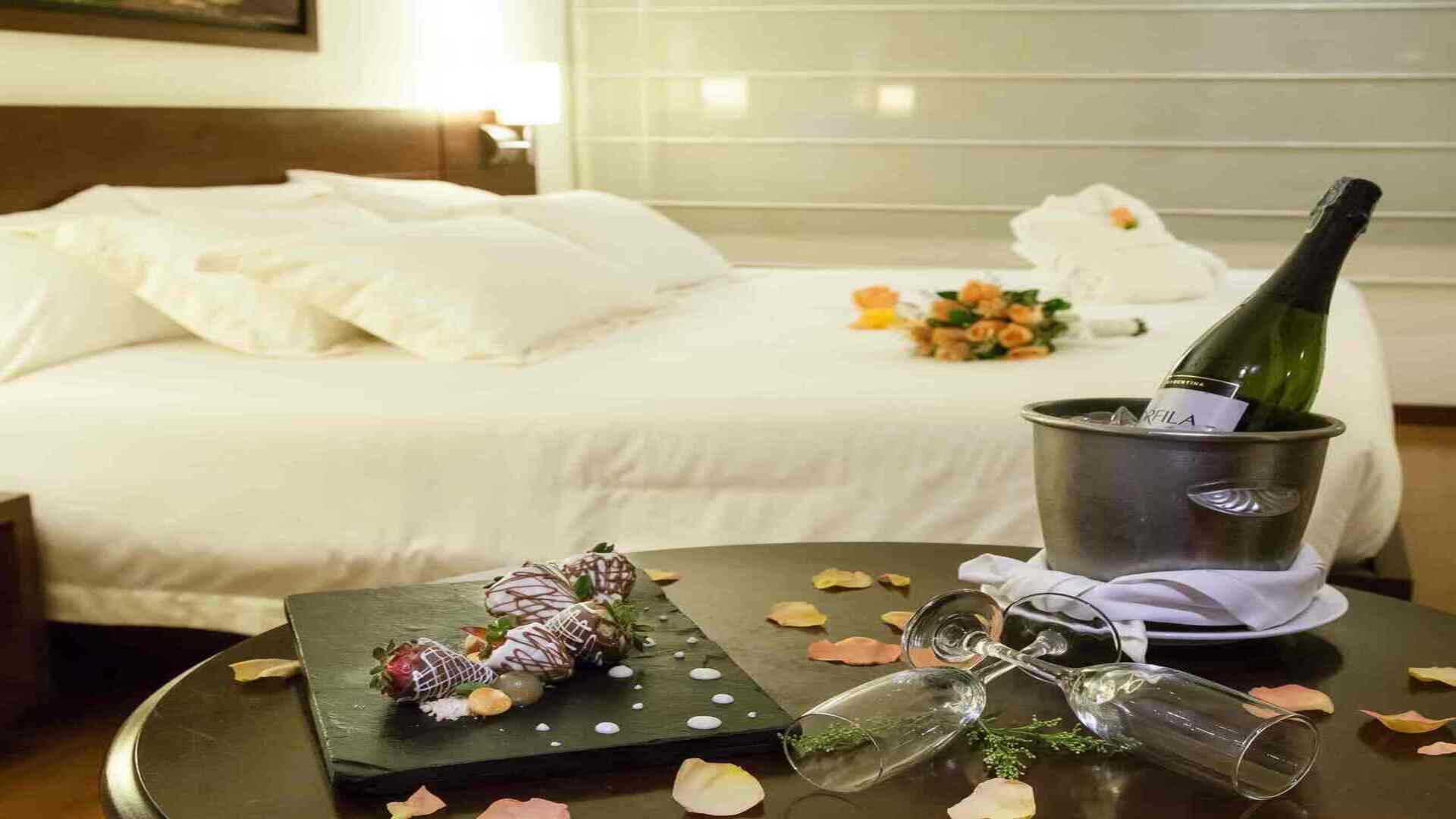 Una cama de hotel decorada para una ocasión especial, con una botella de champán en una cubitera, fresas con chocolate y pétalos de rosa.