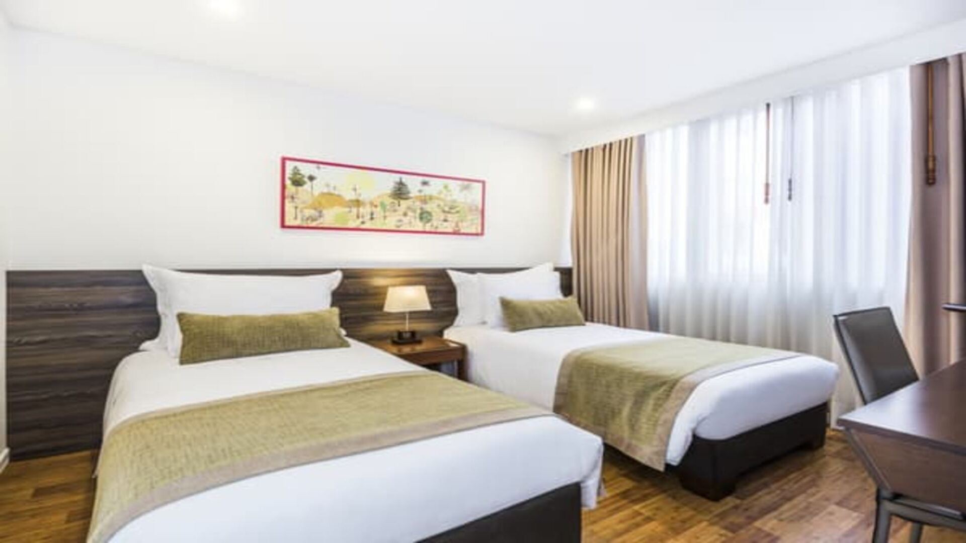 Un dormitorio con dos camas individuales, cada una con ropa de cama blanca y un camino de cama verde. La habitación es luminosa, con una ventana y decoración sencilla.