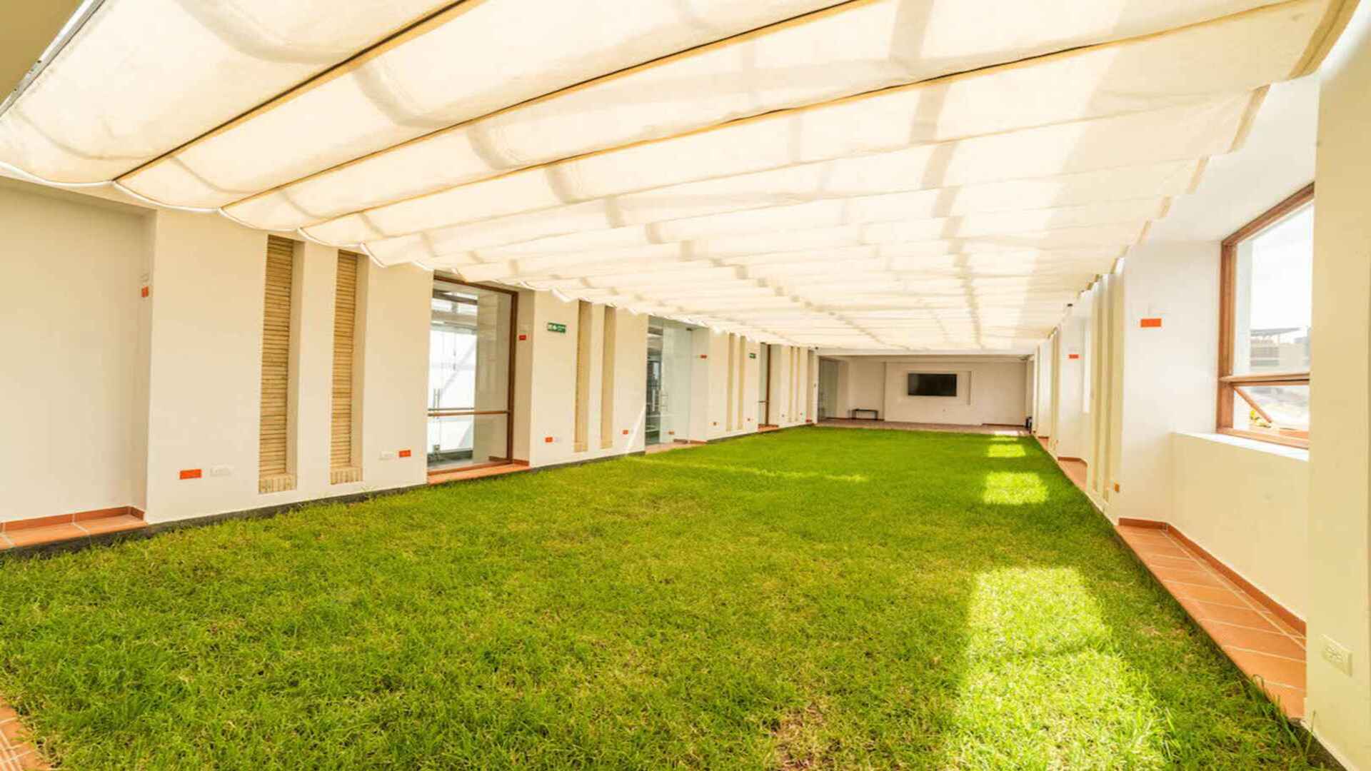  Un amplio espacio interior cubierto con un toldo, que simula un jardín con césped artificial en el suelo. Es un área versátil para eventos o relajación.