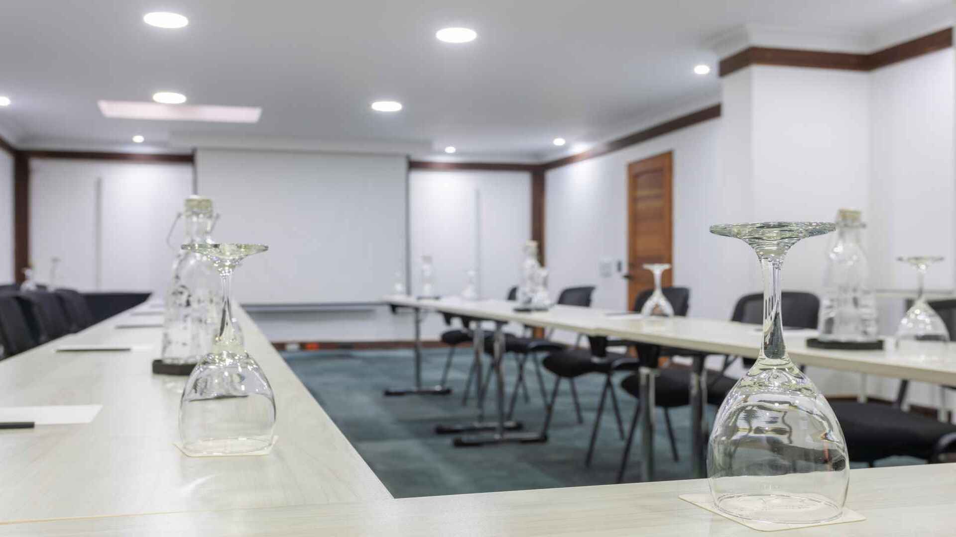 Sala de conferencias preparada con mesas largas y copas invertidas. 